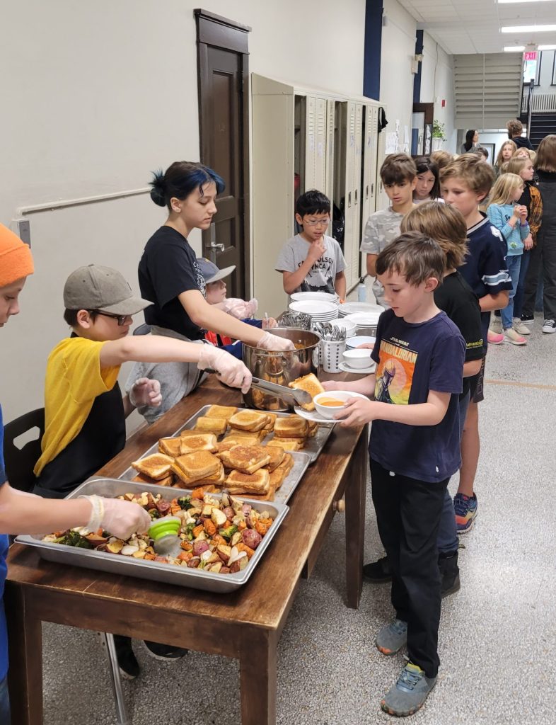 a long line of students wait to receive food being served off a table by four other students