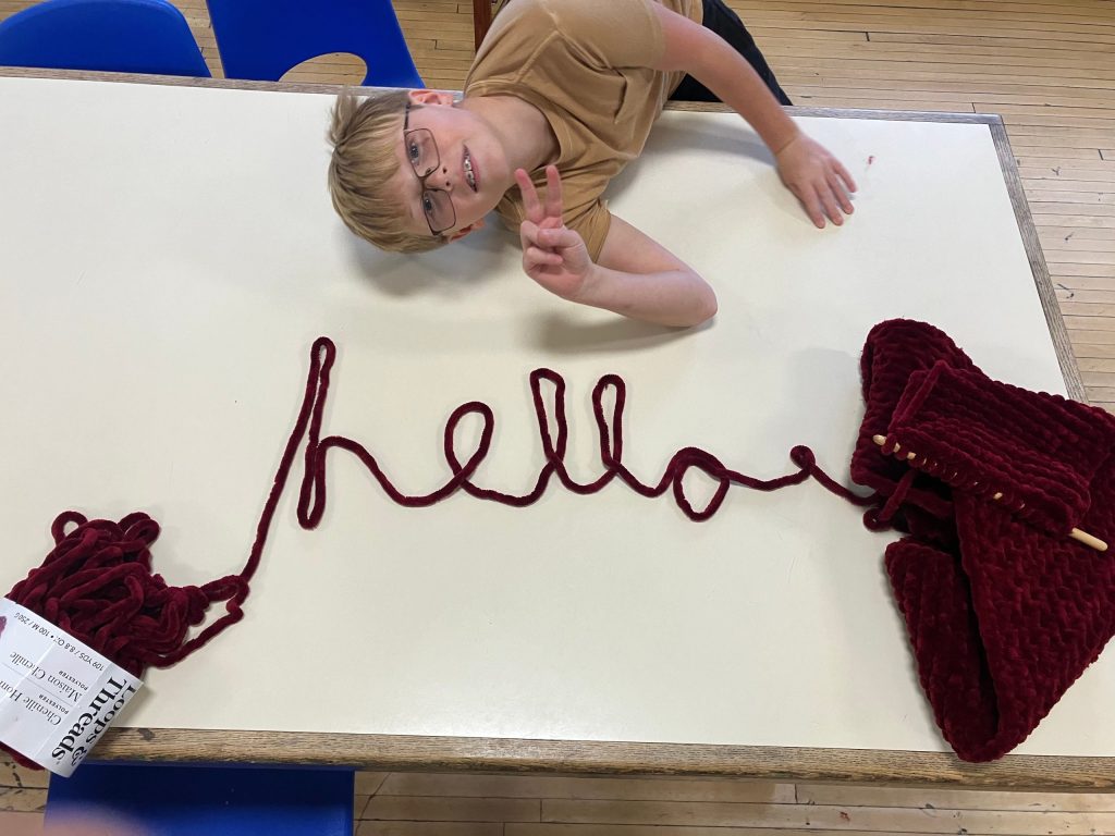 a child gives a peace sign over the word "hello" spelled out in burgundy yarn